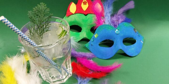 zwei gestaltet Masken in blau und grün auf einem grünem Hintergrund und einem Glas mit Strohhalm und Wacholderzweig